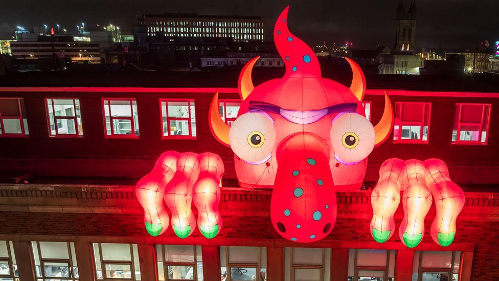 A vibrant red inflatable monster with a long speckled nose and green-nailed fingers peers over the side of an office block in the night-time