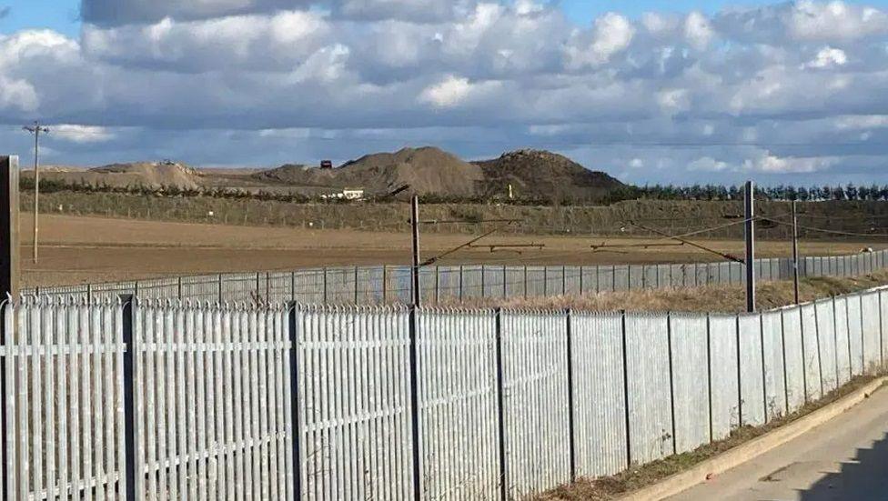 Long metal fences in between a road and brown fields and hills in the background.