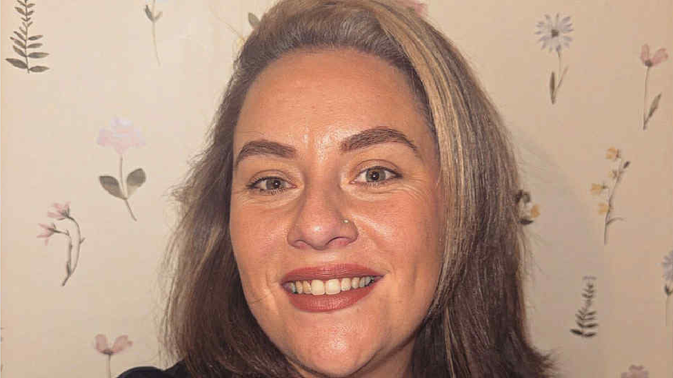 Close-up selfie of Ellie smiling, looking at the camera. She has straight, light brown hair in a bob style; she is pictured standing in front of a wall covered in floral wallpaper.