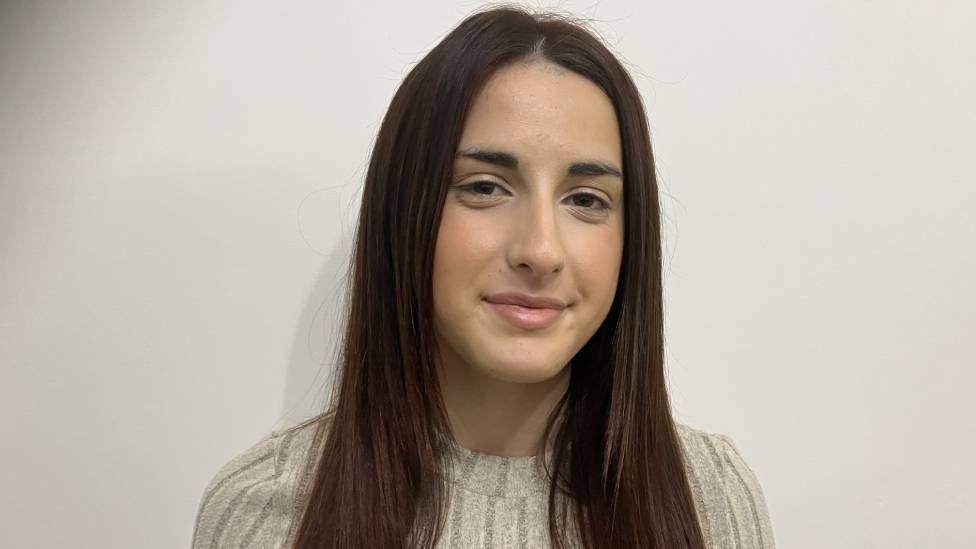 A teenager girl with straight long dark brown hair standing in front of a beige wall 