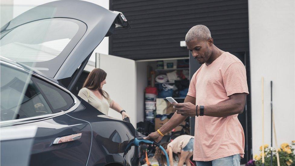 Man using smart phone while charging electric car in front yard