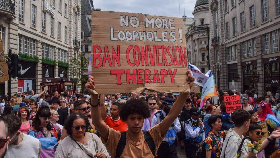 A man in a crowd holds up a sign that says "No more loopholes! Ban conversion therapy"