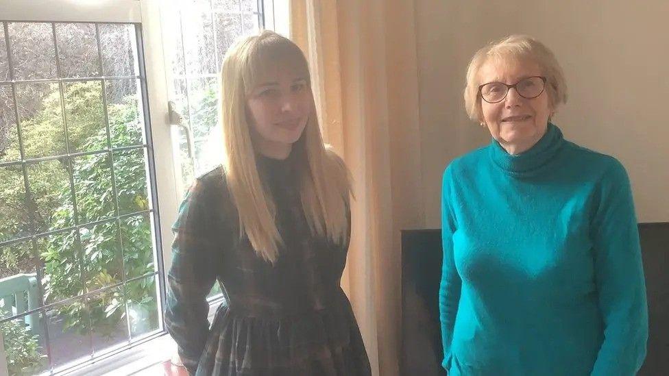Dame Annette at home with one of her Ukrainian lodgers. She has short light red hair, glasses, and wears a blue polo neck jumper. Her friend has long blonde hair and wears a chequered dress.