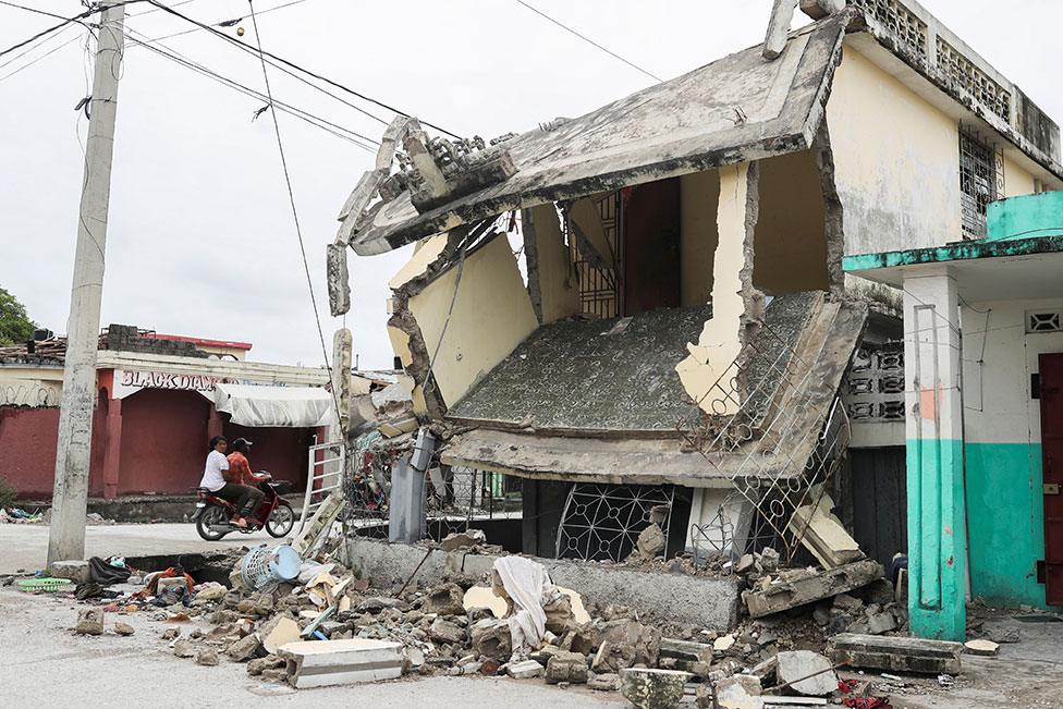 People drive in a motorcycle past a damaged home in Les Cayes, Haiti, on 17 August 2021