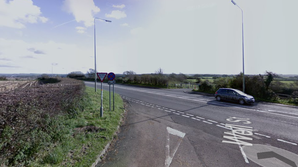 Stretch of road with a car on, next to road signs and near by grass.