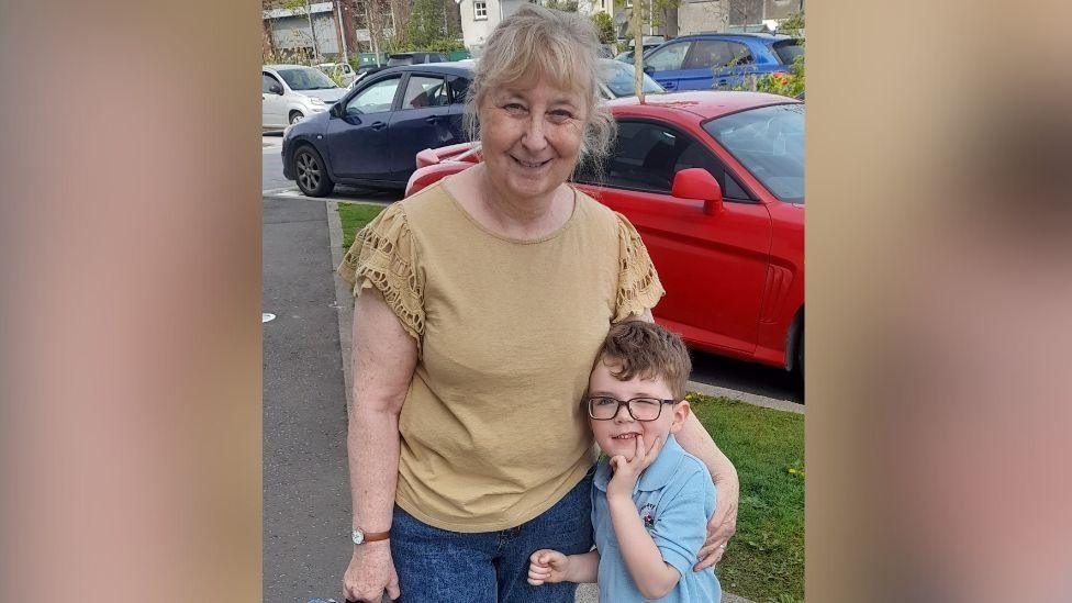 Darlene Garston who has light coloured hair tied back has her arm around her grandson Bertie who is wearing a polo top and has brown hair and glasses