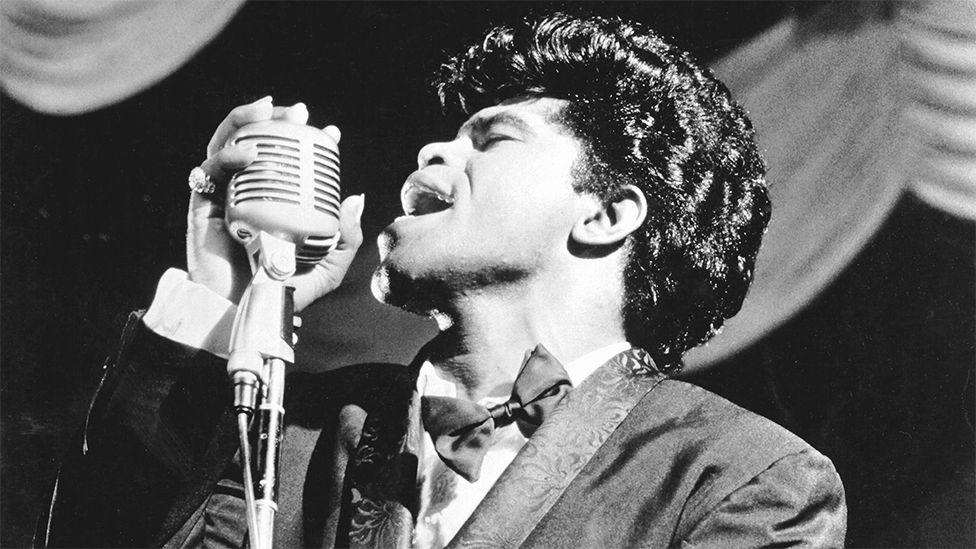 James Brown sings in to a vintage microphone as he performs onstage in 1962 in New York, wearing a suit and bow tie.