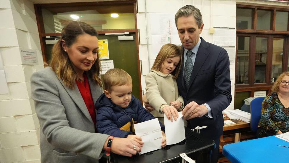 Simon Harris wearing a navy suit holds his daughter, with his wife standing to his right holding their son, as they cast a white ballot into a black voting box