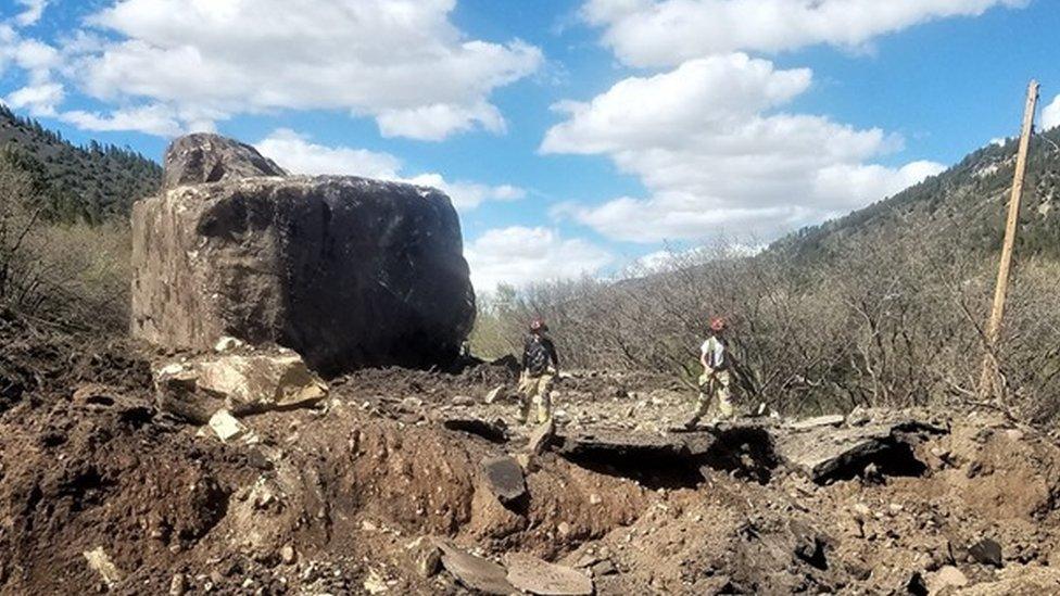 The section of the road that was destroyed by a rock slide containing two enormous boulders with two workers standing in the road.