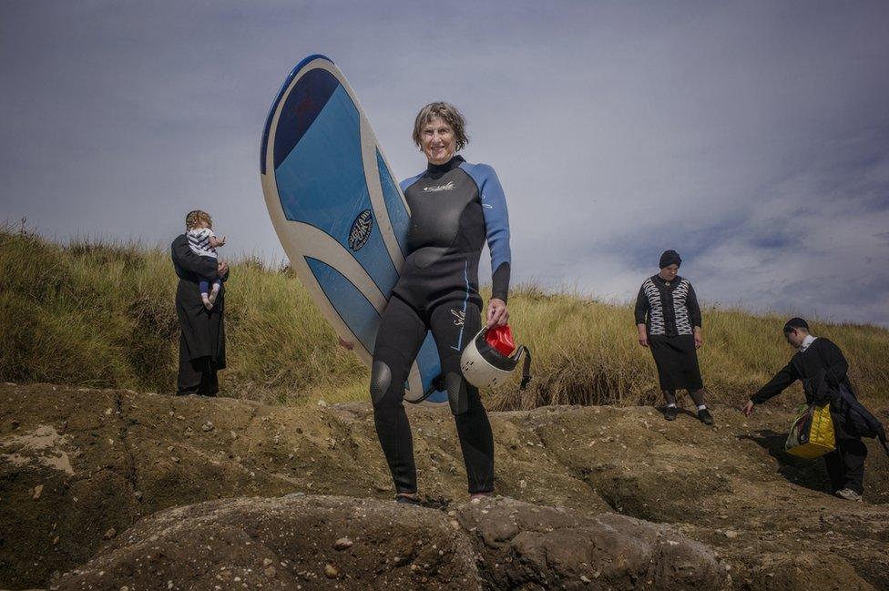 A woman holds a surfboard whilst an Orthodox Jewish family gather behind her