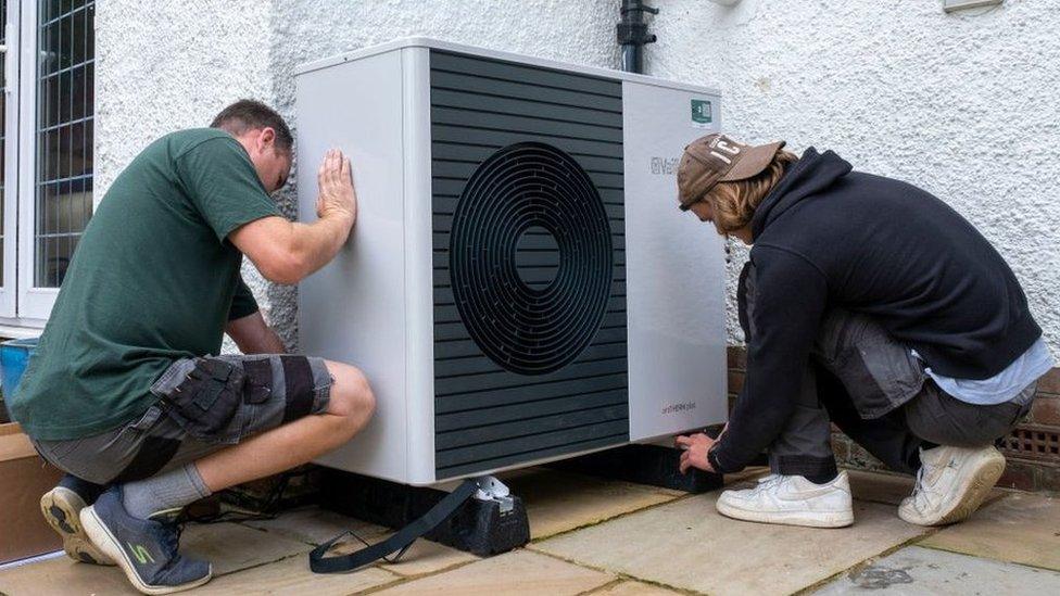 Two men install a heat pump on an external wall of a house in Folkestone. The pump is a large grey box.