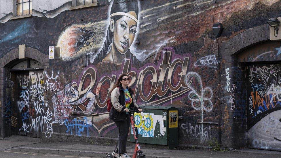 Woman on an eScooter passes the mural on the side of the famous Lakota club in Stokes Croft, one of the original homes of street art and graffiti, on 7th May 2022 in Bristol, United Kingdom.