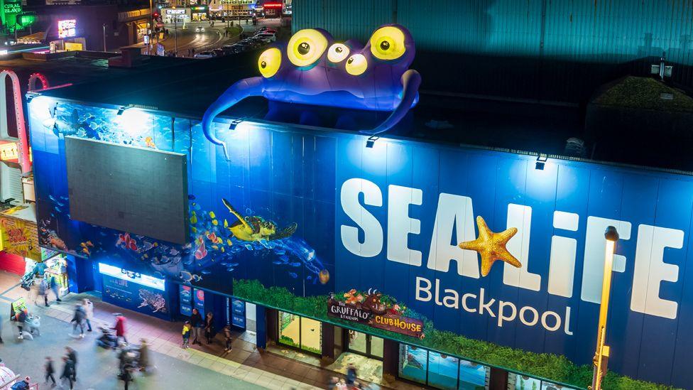 A blue inflatable monster with long tentacles and five eyes peers over the side of the Sea Life centre in Blackpool as people walk by below