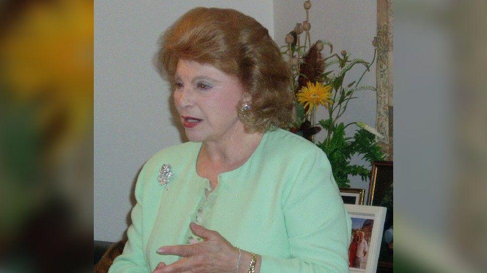 Sally Oppenheim-Barnes speaks in a living room type environment in a picture that may have been taken in the 2000s. She is older, and has big, ginger hair. She is wearing a mint green cardigan with a silver brooch over a mint floral dress, and has red lipstick on. Behind her is a vase of flowers and family photographs in frames.