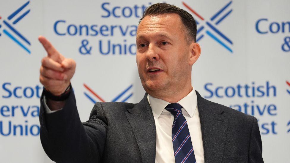 Russell Findlay, with dark hair and a dark suit, pointing in front of Scottish Conservative signage during a press conference 