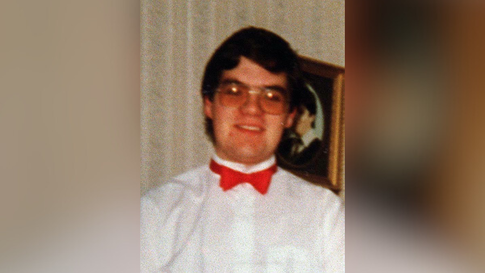 Young man wearing glasses, a white shirt and red bowtie smiling at the camera. He is standing in front of a framed photograph handing on a wall.