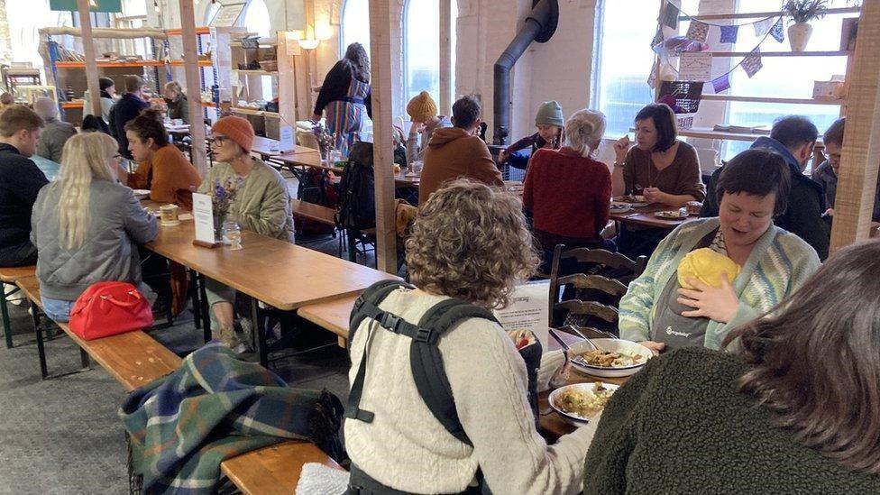 Groups of people are eating meals on benches inside a restaurant run by a charity called The Long Table in Stroud.