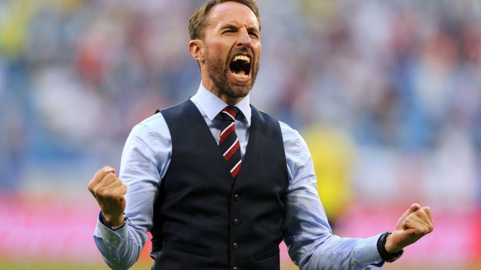 Gareth Southgate, a man in a pale blue suit and navy waistcoat, celebrates with his fists clenched and his mouth wide open on a football pitch
