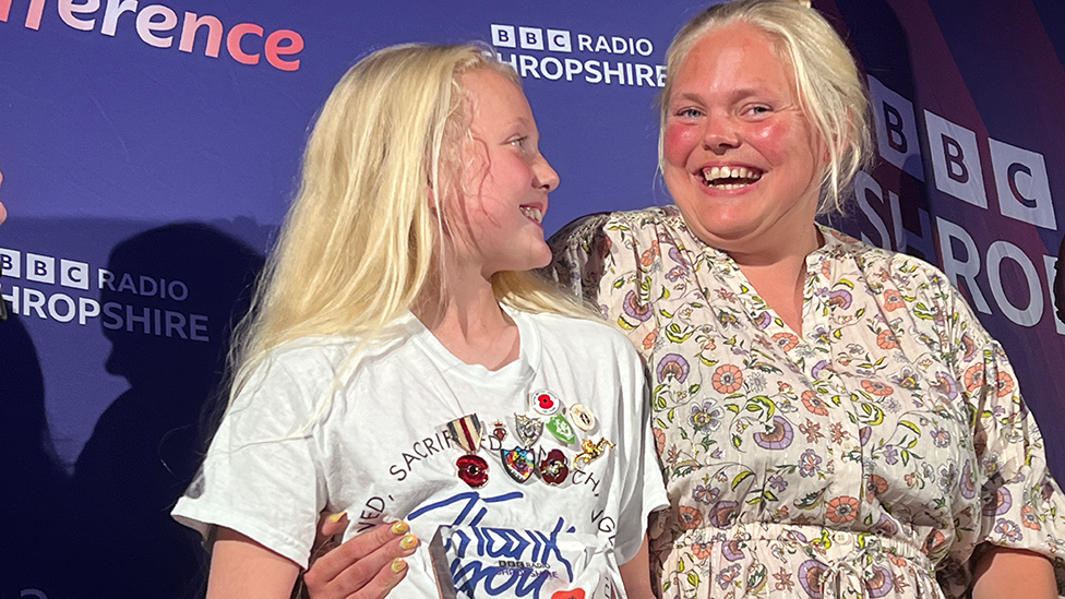 Ruby, wearing a t-shirt covered in Royal British Legion and Blue Peter badges, standing on stage with her mum, picking up her award