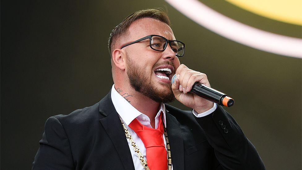 Gareth performs at Leeds Festival in 2022. He is singing into a black microphone, wearing a black blazer, white shirt and red tie.