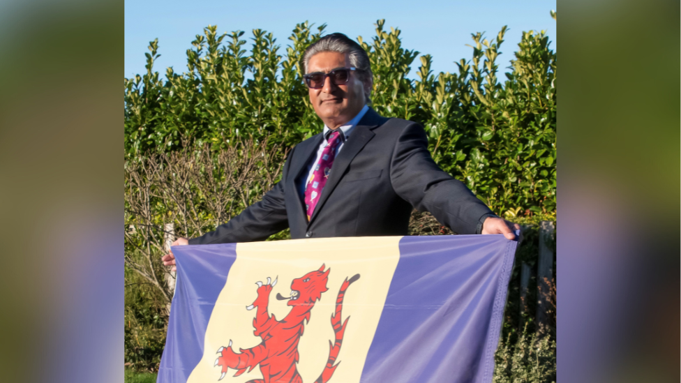 Dal Roy is holding a Fenland flag in both hands while stood outside. He is wearing a suit with a pink tie and is wearing sunglasses. 