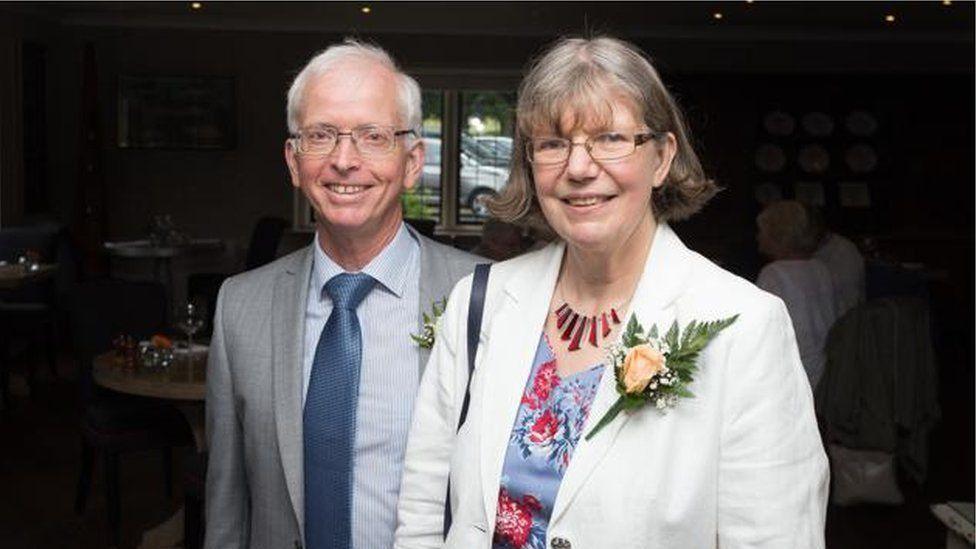 A couple smile at the camera. He is on the left and has short grey hair and glasses, and is wearing a grey suit with a blue short and tie. She is on the right, has short greying hair in a bob, glasses and is wearing a blue and red floral dress and a white jacket with a flower attached to the chest