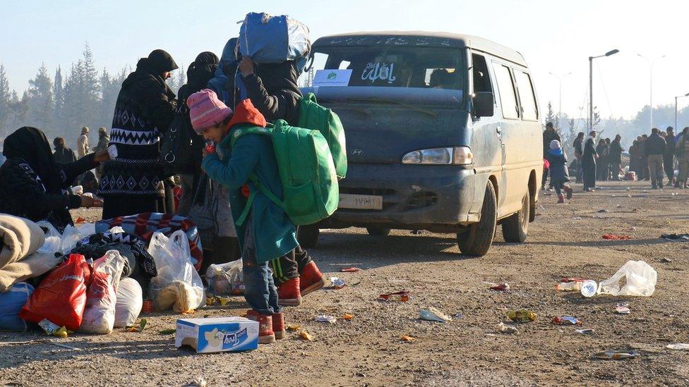 A child wearing a large backpack stands next to piles of the family's belongings