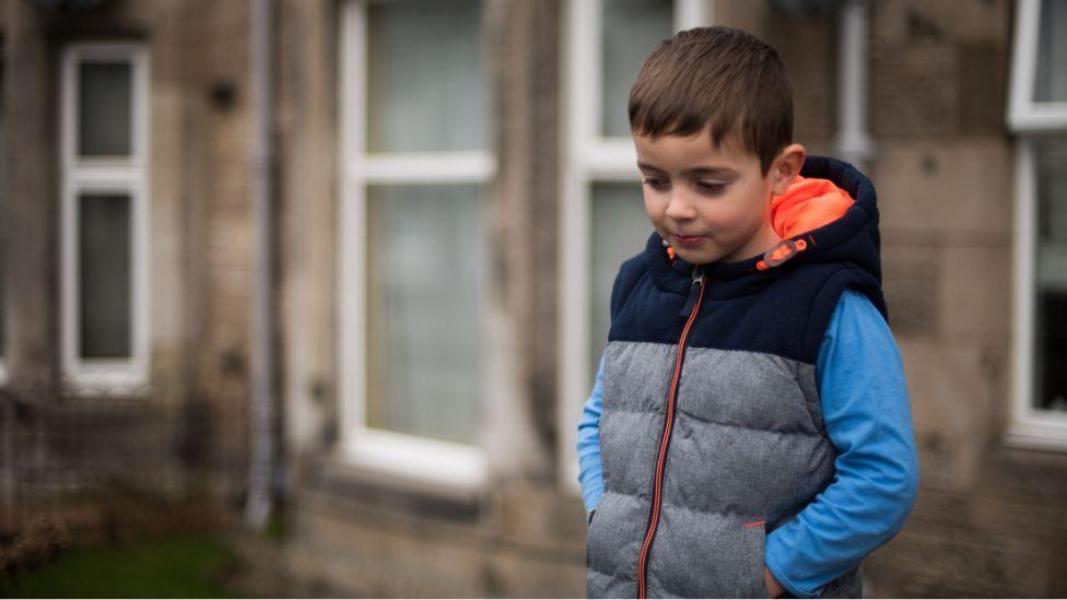 A sad-looking child with dark hair, wearing a grey body warmer over a blue top, looks at the ground while standing in a street 