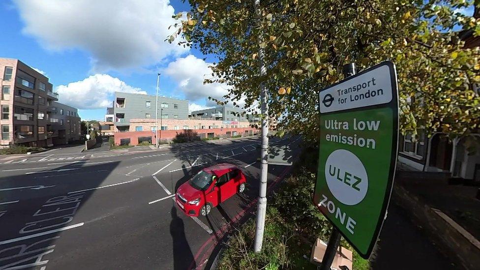 A ULEZ sign next to a red car on a road in London.