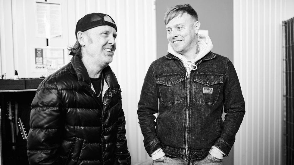 A black and white photo of Sam Carter and Metallica drummer Lars Ulrich. Sam's standing with his hands in his pockets, looking fondly at Lars. Lars is perched on some sort of box or equipment, and smiling. He's wearing a black puffer jacket and backwards baseball cap. They appear to be in a backstage area and the mood is relaxed.