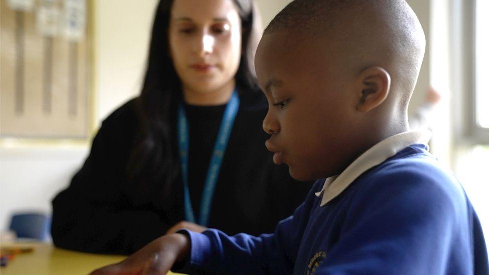 A boy in a side profile of a blue school uniform jumper with short hair sits at a table. Behind him sits a teacher with a black top, blue lanyard and long brown hair looking at his work. 