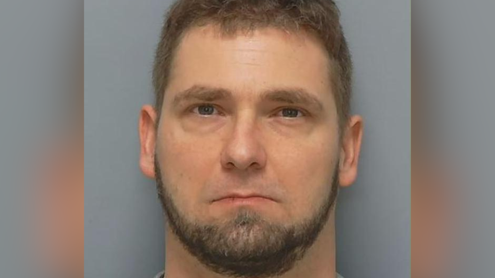 A custody picture of a white man with brown hair and brown beard.