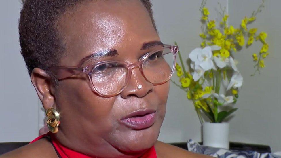 Edith Nyachuru, seated and wearing a red top, reading glasses and gold earrings. A vase of flowers can be seen behind her - 13 November, Zimbabwe