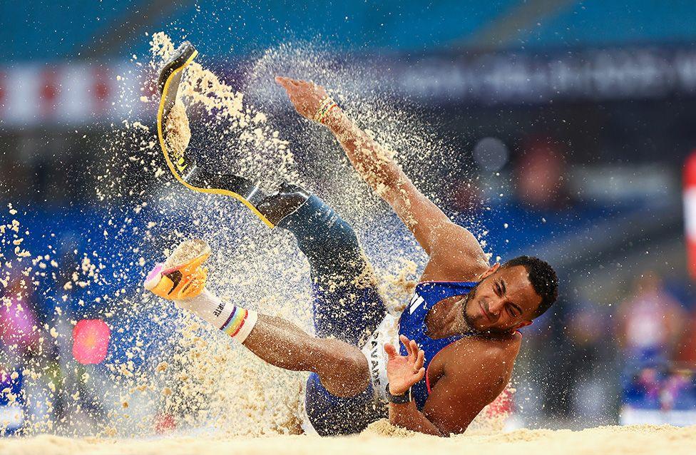 Dimitri Pavade competes in the Men’s Long Jump F64 Final at the World Para Athletics Championships in New Delhi. He lands in a sandpit wearing a blue uniform and a curved carbon-fiber prosthetic blade, with sand spraying upward against a stadium backdrop.