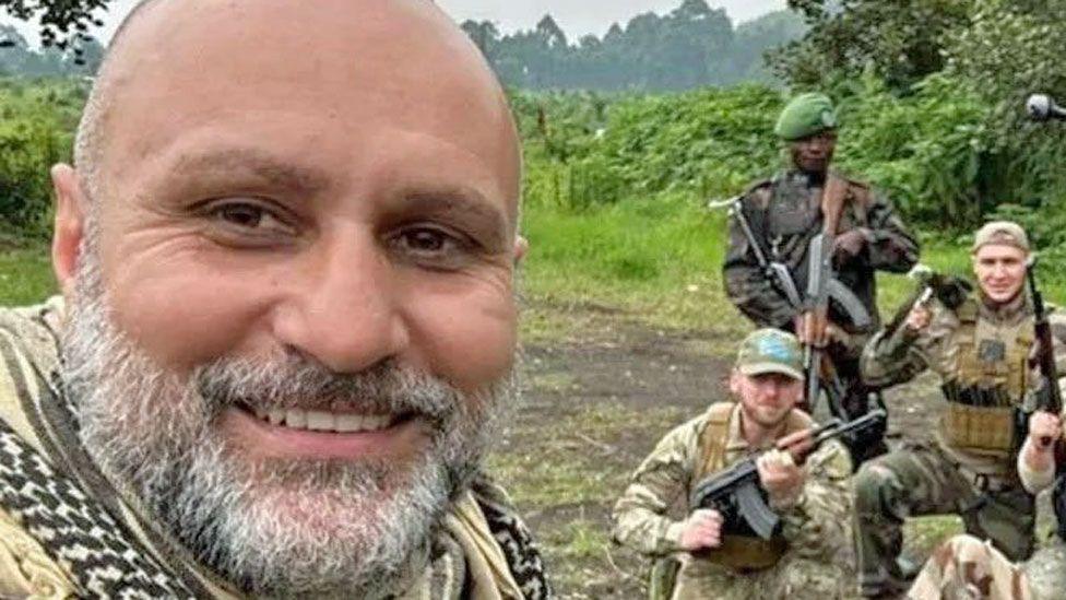 Vasile Badea, taking a selfie whilst in DR Congo. He has a grey beard, closely cropped, nearly bald head and a chequered scarf. Behind him to the left can be seen other a few other mercenaries along with a member of the Congolese armed forces - all posing with guns.