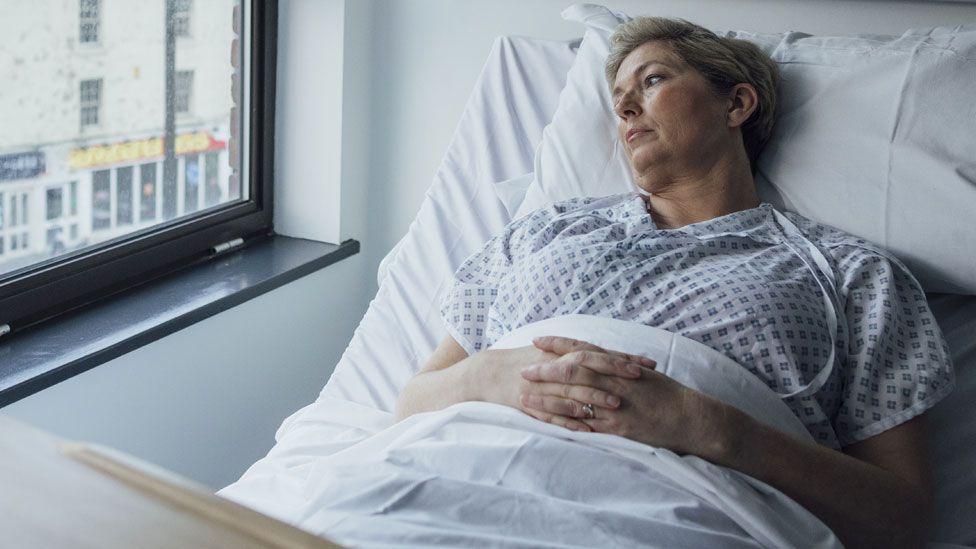 A woman lies in a hospital bed and looks out of a window