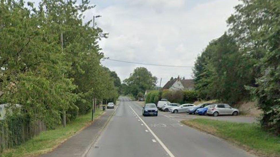 A view of a straight section of the road with trees, one car driving and some parked off street at the side