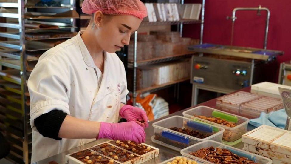 Chocolate worker in white overall and hair net and latex gloves adds nuts to bars of chocolate in a chocolate factory setting