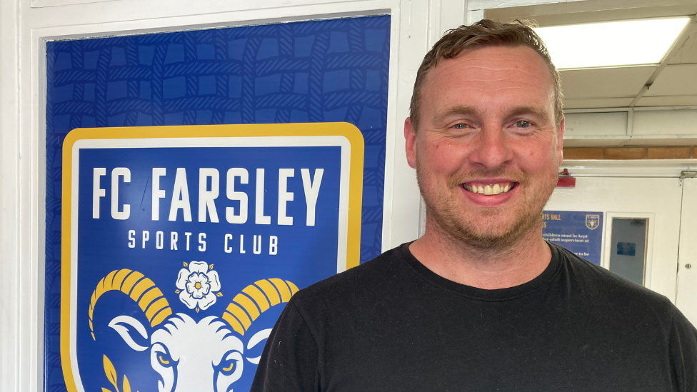 David Stockdale is wearing a black t-shirt and is stood in a doorway beside a coat of arms style sign with Farsley FC written on it