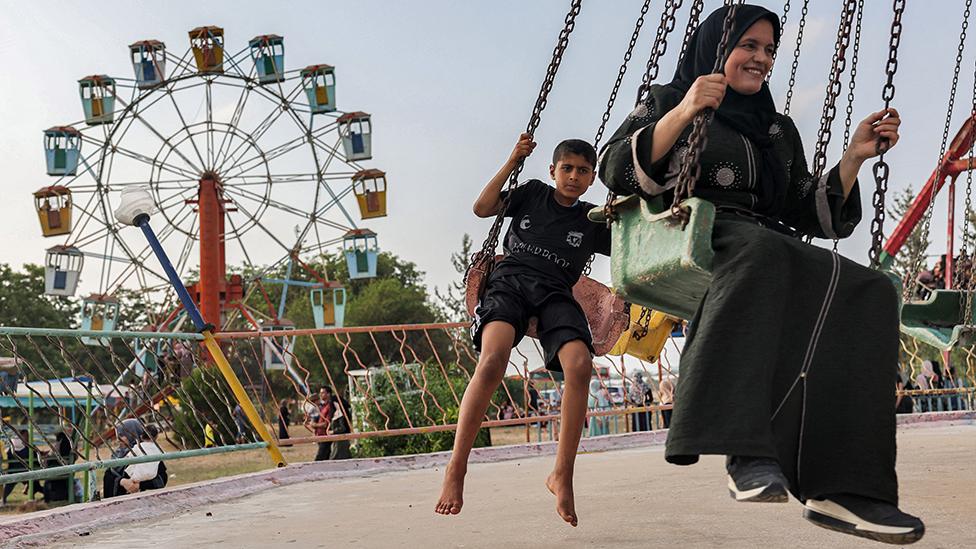 A Palestinian woman dressed in black wearing a headscarf laughing on a swing ride at the park in 2022 with a boy in dark shorts on the seat behind her - the ferris wheel with red, blue, yellow and green passenger cars can be seen in the background