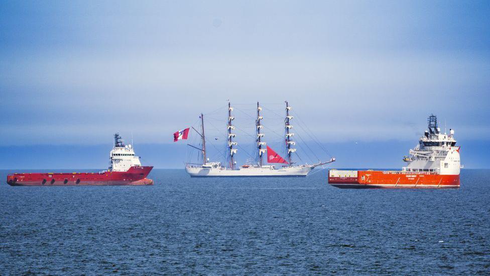 Tall ship BAP Union, flanked by two offshore industry vessels, under a blue sky.