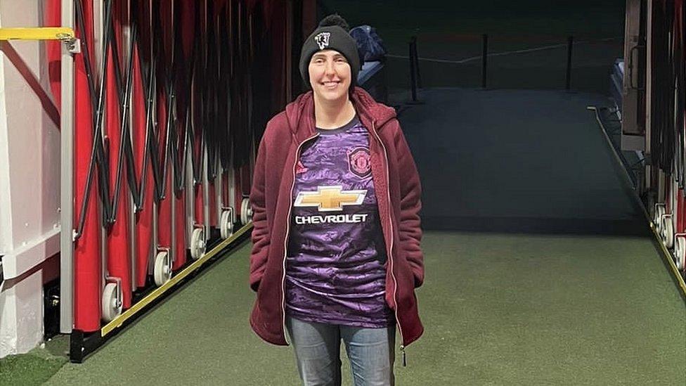 Zoe wearing a purple Manchester United shirt, a burgundy hoodie, black beanie and jeans facing the camera while stood in the tunnel at Manchester United's ground, Old Trafford.