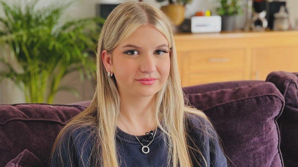 Keira sitting on a purple sofa with a big green plant in the background and a brown wooden unit. She has blonde hair and has her nose and ears pierced. She has a ring on a necklace around her neck and has a purple jumper on.