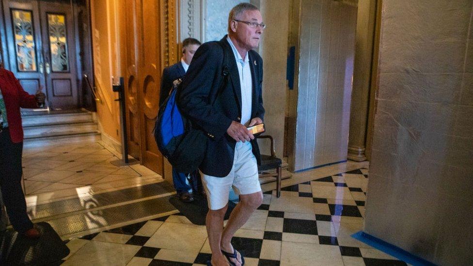 Man wearing a blazer, shorts and flip flops walks on black and white marble floor in front of a man in a suit.