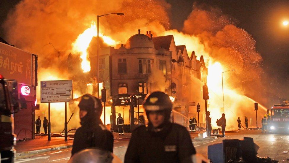 Two police officers in riot gear walk in front of a burning building in London