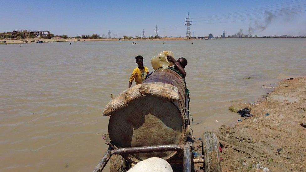 Two boys fill drum up on a cart, with water from buckets from the river