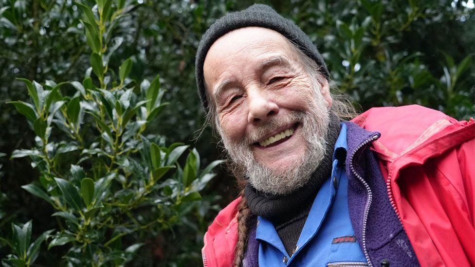 Bob Flowerdew standing in front of an evergreen hedge. He has a grey beard and a plait of hair over his left shoulder. He is wearing a grey woolly hat and a red waterproof over a purple fleece, a blue shirt and a black roll-neck jumper and is smiling broadly