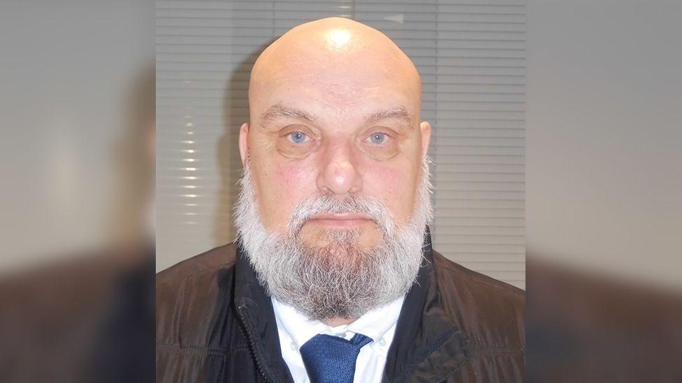 Custody photograph of Hedley Heddison, who is bald with a large white beard and wearing a brown jacket with white shirt and blue tie.