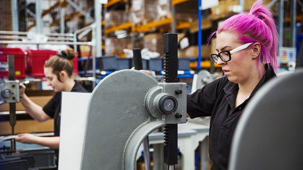 A woman with pink hair uses a hole punch machine in a factory making bicycles, with another worker in the background in a manufacturing environment.