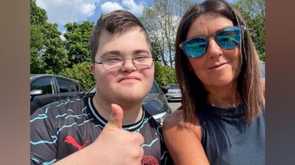 A boy in a black and blue stripy t-shirt with glasses and his thumb up next to a woman in a dark blue top and reflective sunglasses